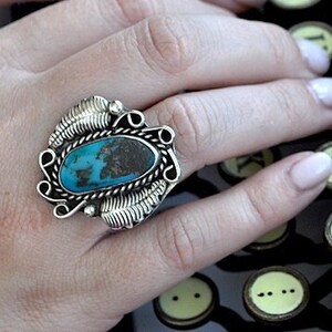 May include: A silver ring with a turquoise stone set in a decorative design. The ring is worn on a person's finger as they type on an antique typewriter labeled "CORONA".