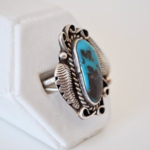 May include: A silver ring with a turquoise stone set in a detailed design. The ring is on a white display stand.