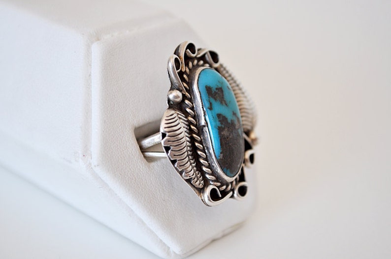 May include: A silver ring with a turquoise stone set in a detailed design. The ring is on a white display stand.