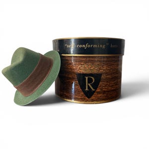 May include: A green fedora hat with a brown band rests beside a cylindrical hat box. The hat box has a wood-grain pattern and a black band with the text "self-conforming hats". A gold "R" is on the front.