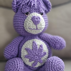 May include: Purple crocheted teddy bear with a gray crocheted marijuana leaf on its chest. The bear has a white crocheted nose and mouth.