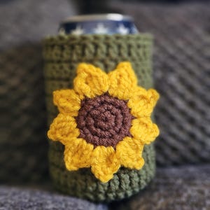 May include: A green crochet can cozy with a yellow and brown sunflower applique. The cozy is designed to fit a standard-sized can. The sunflower has a brown center and yellow petals. The cozy is made of a soft, textured yarn.