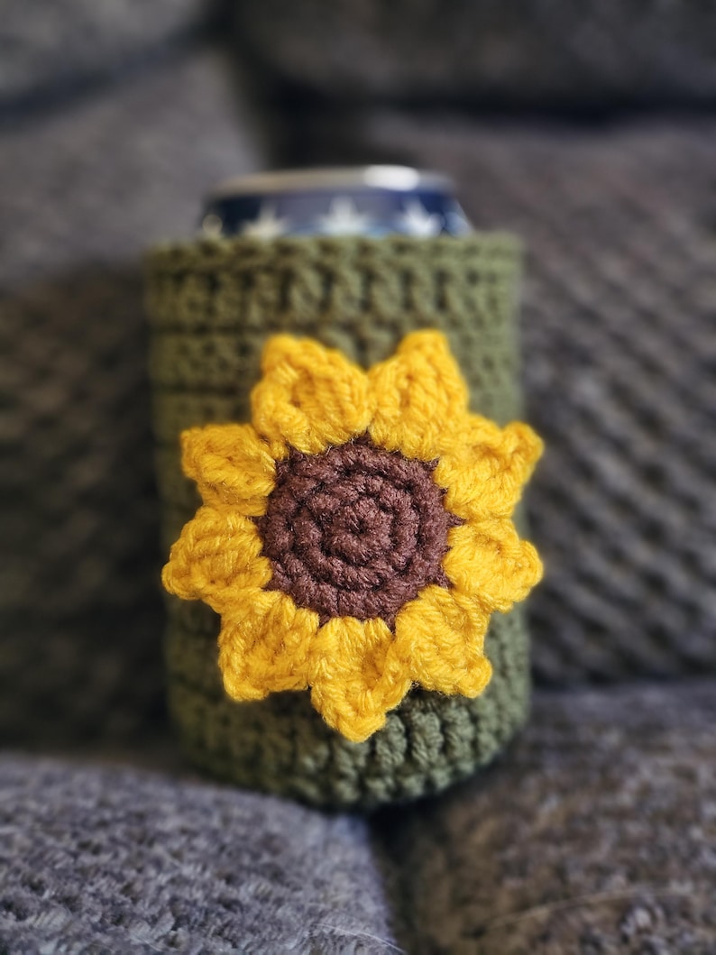 May include: A green crochet can cozy with a yellow and brown sunflower applique. The cozy is designed to fit a standard-sized can. The sunflower has a brown center and yellow petals. The cozy is made of a soft, textured yarn.