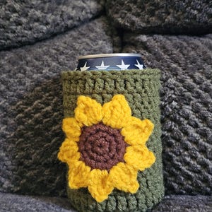 May include: A green crochet can cozy with a large yellow and brown sunflower design. The cozy is wrapped around a can with a blue and white star pattern. The cozy is sitting on a gray, textured surface.