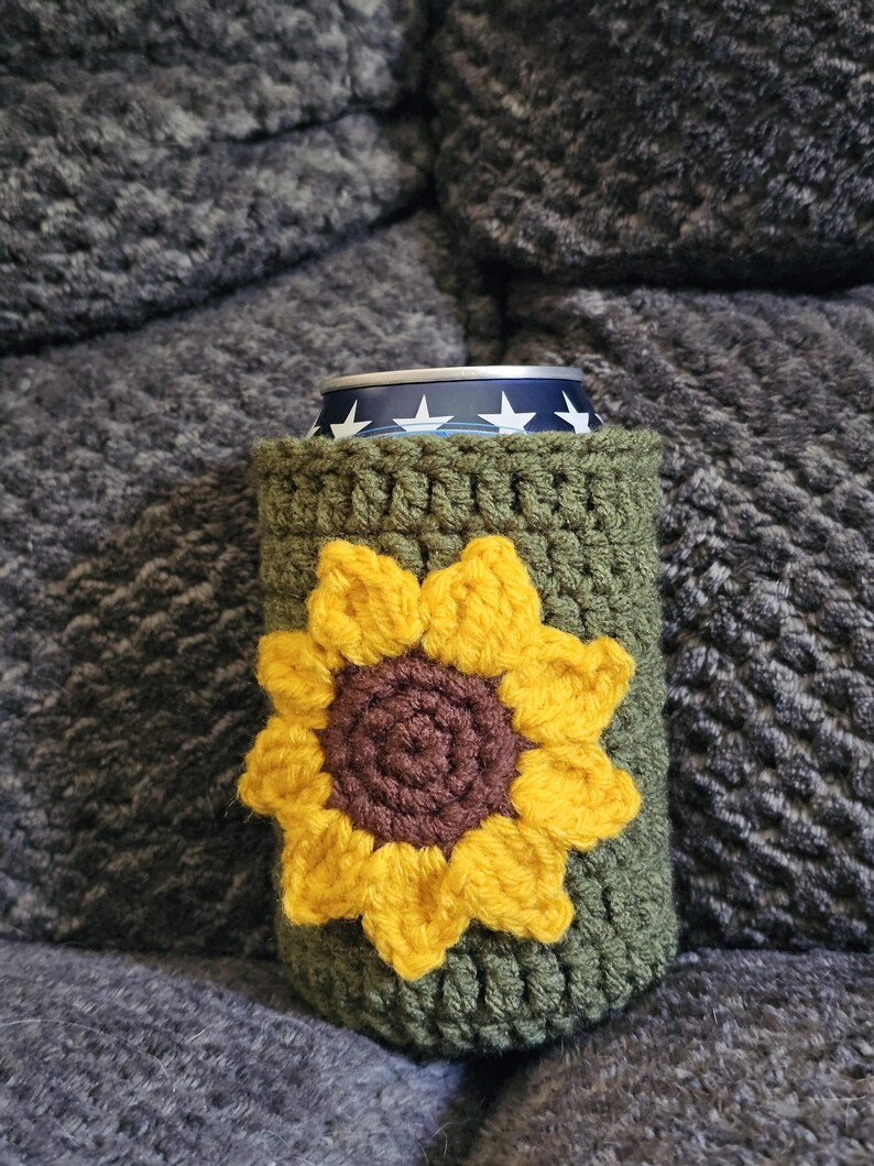 May include: A green crochet can cozy with a large yellow and brown sunflower design. The cozy is wrapped around a can with a blue and white star pattern. The cozy is sitting on a gray, textured surface.