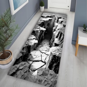 Op de afbeelding: Een zwart-wit rechthoekig tapijt met een 3D-canyonontwerp. Het tapijt heeft een gedetailleerde weergave van een diepe canyon met gebarsten aarde en torenhoge rotsformaties. Het ontwerp creëert een optische illusie van diepte.