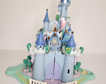 May include: A light blue and purple toy castle with multiple turrets, a clock tower, and a pink staircase. The castle has a light green base with a white railing and small green trees. The castle doors are purple.
