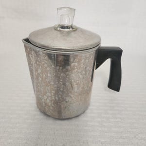 May include: A vintage aluminum coffee percolator with a clear glass knob on the lid and a black handle. The metal has a speckled appearance. The percolator is designed for brewing coffee.