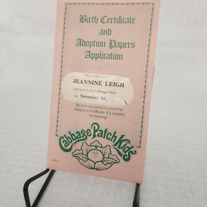 May include: A pink Cabbage Patch Kids birth certificate and adoption papers application, displayed on a black metal stand. The certificate includes the name "JEANNINE LEIGH" and the date "November 1st". The Cabbage Patch Kids logo is visible.