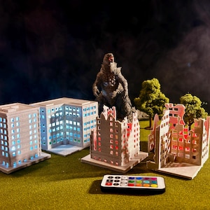 May include: A miniature city scene with a Godzilla figure, several building models, and a remote control. The buildings are lit with blue and red lights, suggesting a city under attack. The scene is set on a green surface.