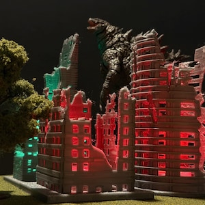 May include: A miniature diorama depicting a city in ruins, with several buildings illuminated by red and green lights. A Godzilla figure looms in the background, and a small tree is in the foreground. The scene evokes a sense of destruction and fantasy.