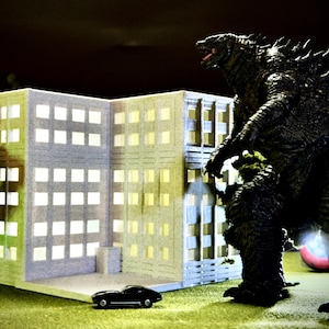 May include: A miniature cityscape scene featuring a towering Godzilla figure, a multi-story building with lit windows, a small black car, and a green grassy area. The dark background adds to the dramatic effect.