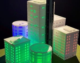 May include: Five white 3D printed buildings with different designs and sizes. The buildings are lit from within with green, blue, and red lights. The buildings are on a green surface.