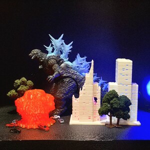 May include: A Godzilla figure with blue dorsal fins is positioned amongst miniature buildings and trees. An orange explosion and toy cars are also present. The figure is dark grey and blue, set against a dark background.