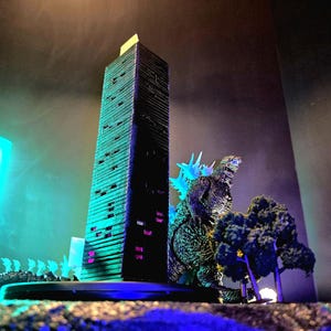 May include: A miniature Godzilla figure stands near a tall, dark skyscraper model. The scene includes other miniature buildings and trees, all illuminated with blue and green lighting, creating a dramatic effect. The overall theme is a city under attack.
