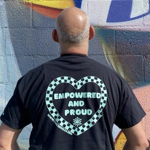 May include: Black t-shirt with a heart-shaped design in a checkerboard pattern. The text "EMPOWERED AND PROUD" is inside the heart. The website address www.tascservices.org is below the heart.