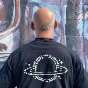 May include: Black t-shirt with a white graphic of a planet with rings and stars. The text on the shirt reads "THE ADULT SKILLS CENTER" above the planet and "CHANGING THE UNIVERSE" below it. The background is a colorful mural.