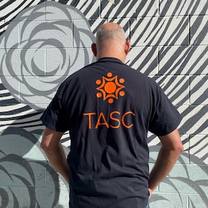 May include: Black t-shirt with the orange text "TASC" and an orange graphic on the back. The person is standing in front of a mural with a black, white, and gray design.