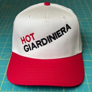 May include: White baseball cap with a red brim. The front of the cap has black embroidery that reads "HOT GIARDINIERA".