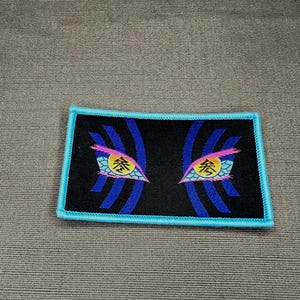 May include: A rectangular embroidered patch with a black background and a light blue border. The design features two stylized eyes with yellow irises, each containing the black Japanese kanji character for three, surrounded by blue and pink accents.