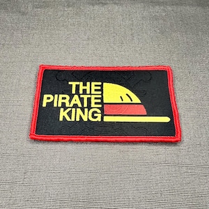 May include: A rectangular embroidered patch with a black background and a red border. The text THE PIRATE KING is stitched in yellow, accompanied by a yellow and red graphic of a hat. A faint, embossed skull and crossbones design is visible.