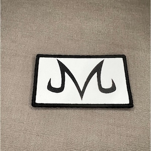 May include: A rectangular embroidered patch featuring a white background with a black border and a stylized black letter M symbol centered on the surface. The design is clean and bold, with a thick black stitched edge framing the white fabric.