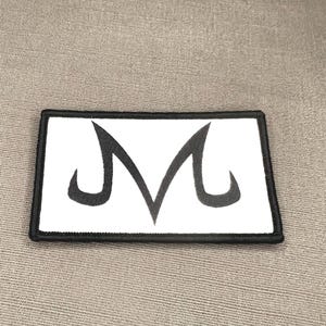 May include: A rectangular embroidered patch featuring a white background with a bold black border. Centered on the patch is a stylized black letter M symbol with sharp, curved edges, characteristic of the Majin mark from Dragon Ball Z.