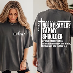 May include: Dark gray t-shirt with white text. The front says "LET'S PRAY" with a cross. The back says "NEED PRAYER? TAP MY SHOULDER" with a cross and stars, and a Bible verse. A casual, faith-based apparel item.