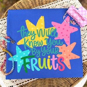 May include: A royal blue t-shirt with the phrase "They Will Know You By Your Fruits" in colorful lettering. The shirt features star-shaped graphics in yellow, pink, orange, and turquoise. A pink and white hair clip and beaded necklaces are also visible.