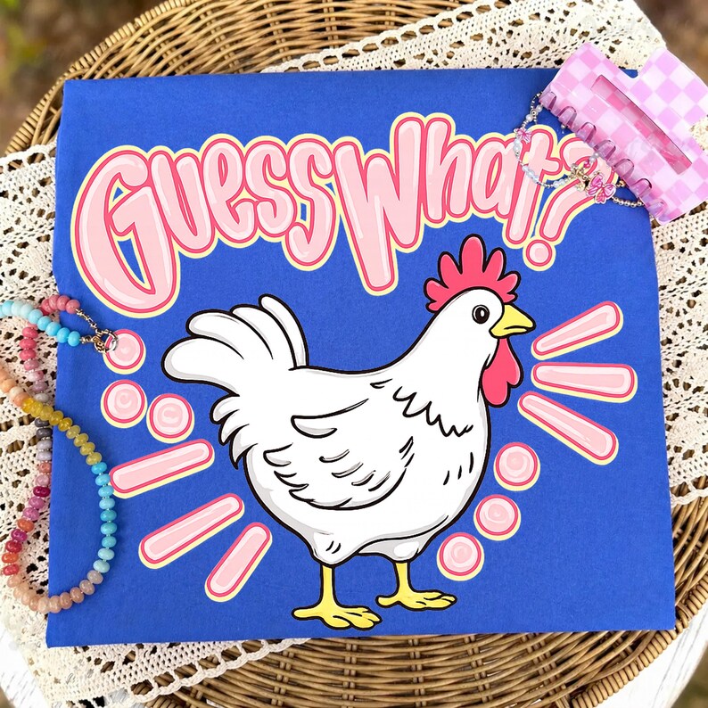 Puede incluir: Una camiseta azul con el texto "Guess What?!" en letras rosas con burbujas. Un dibujo de un pollo blanco con cresta roja y pico amarillo aparece debajo del texto. Tambi&eacute;n se ven una pinza para el pelo rosa y un collar de cuentas.