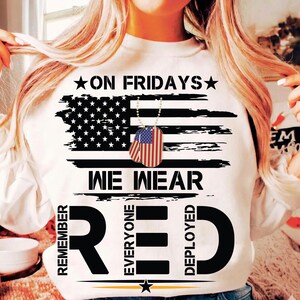 May include: White sweatshirt featuring a distressed American flag design. The text reads "ON FRIDAYS WE WEAR RED" with the words "REMEMBER EVERYONE DEPLOYED" around the word RED. Includes a dog tag necklace.