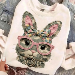 May include: White sweatshirt featuring a detailed applique of a bunny face with pink glasses, a bow, and a rose. The bunny design incorporates camouflage fabric and pink accents. The sweatshirt is displayed on a denim jacket.