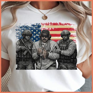 May include: White t-shirt featuring a graphic of three soldiers in military gear, standing in front of a distressed American flag. The soldiers are in shades of gray and black, with the words "U.S. NAVY" visible on their uniforms.