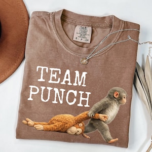 Op de afbeelding: Bruin t-shirt met de witte tekst "TEAM PUNCH". Het shirt heeft een afbeelding van twee apen, een liggend en de ander lopend. Een zilveren ketting met een hanger is ook zichtbaar.