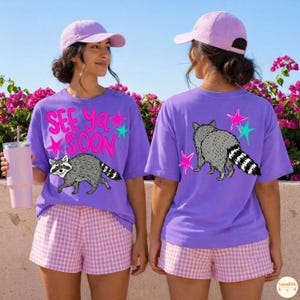 May include: Purple t-shirts with a raccoon graphic and the text "See Ya Soon". The front of the shirt features a raccoon illustration and the text in pink and teal. The back of the shirt has a raccoon illustration and pink and teal stars. The model is wearing a pink baseball cap and pink and white checkered shorts.