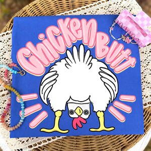 Puede incluir: Una camiseta azul con la frase "CHICKEN BUTT" en letras rosas con burbujas y una ilustraci&oacute;n de un pollo de dibujos animados. El pollo est&aacute; al rev&eacute;s, con la parte trasera mirando al espectador. Tambi&eacute;n se ven un collar y una pinza para el pelo.