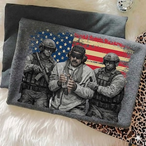 May include: Gray t-shirt featuring a graphic of three U.S. Navy soldiers with an American flag backdrop. The design includes detailed illustrations of the soldiers in uniform, holding weapons and equipment. The flag design has a distressed look.
