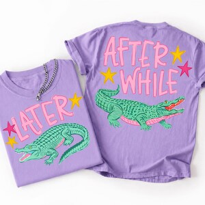Puede incluir: Dos camisetas moradas con un diseño de cocodrilo verde. Una camiseta dice "LATER" y la otra "AFTER WHILE" en letras rosas. Ambas camisetas tienen detalles de estrellas amarillas y rosas. Una cadena de plata está en una camiseta.