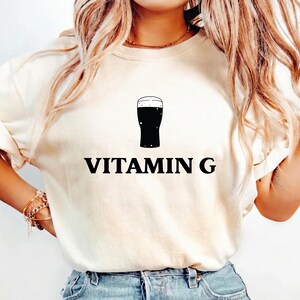 以下が含まれることがあります： 黒い文字で「VITAMIN G」と書かれたクリーム色のTシャツ。その下に黒いビールのパイントのグラフィックがあります。このシャツはカジュアルなスタイルで、普段使いに適しています。