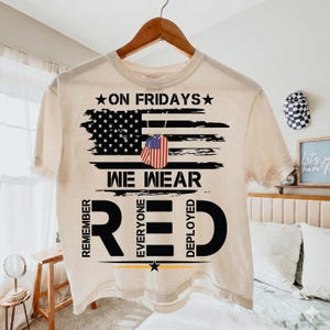 May include: Cream-coloured t-shirt with a patriotic design. The shirt features a distressed American flag graphic with the text "ON FRIDAYS WE WEAR RED" and the words "REMEMBER EVERYONE DEPLOYED".