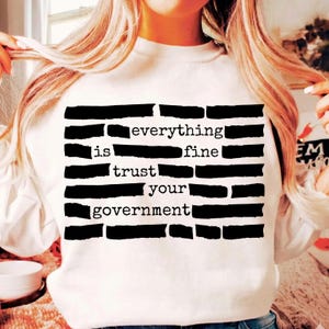 Può includere: Felpa bianca con la scritta "everything is fine trust your government" in nero, in stile bloccato. Il testo è disposto sulla parte anteriore del capo.
