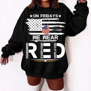 May include: Black sweatshirt featuring the text "ON FRIDAYS WE WEAR RED" and an American flag design. The design includes a dog tag with the American flag. The words "REMEMBER EVERYONE DEPLOYED" are also on the sweatshirt.