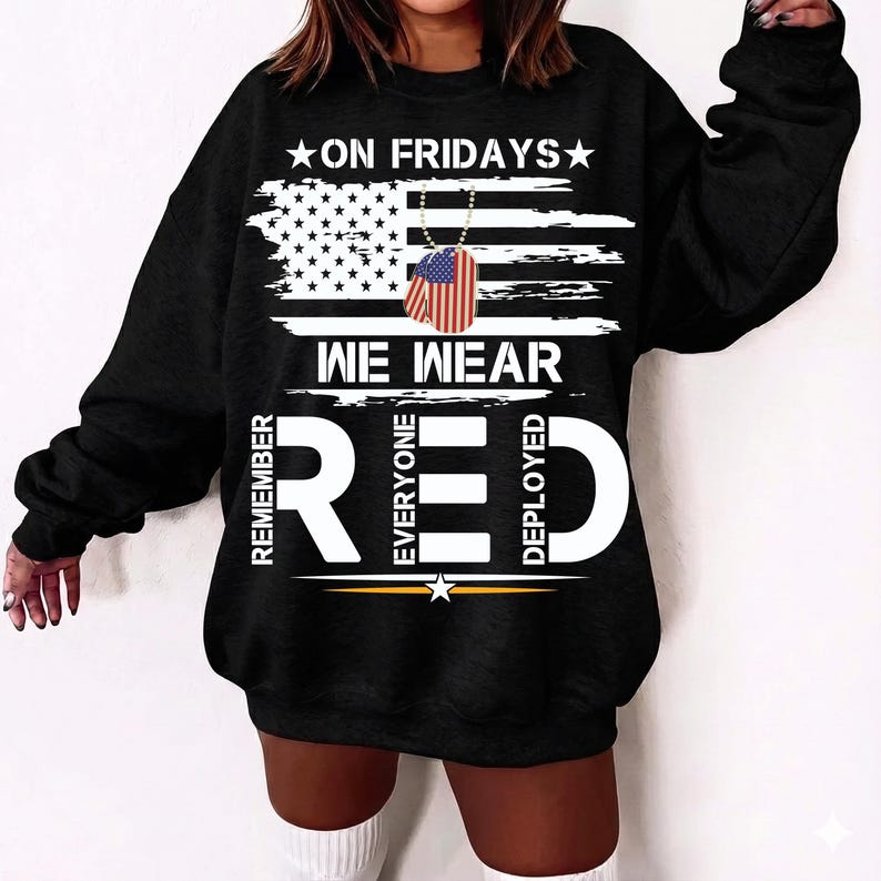 May include: Black sweatshirt featuring the text "ON FRIDAYS WE WEAR RED" and an American flag design. The design includes a dog tag with the American flag. The words "REMEMBER EVERYONE DEPLOYED" are also on the sweatshirt.