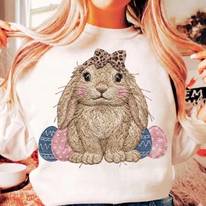 Peut inclure: Sweat-shirt blanc avec une illustration détaillée d'un lapin portant un nœud léopard et des joues roses. Trois œufs de Pâques décoratifs en denim bleu et motifs floraux roses sont en dessous du lapin. Le motif est centré sur le devant du t-shirt.