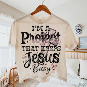 Puede incluir: Camiseta color crema con el texto "I'M A Project THAT KEEPS Jesus Busy" en letras negras y rosas. La camiseta est&aacute; colgada en una percha de madera.