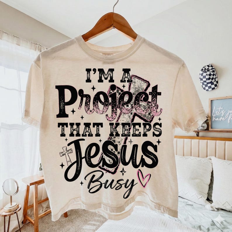 Puede incluir: Camiseta color crema con el texto "I'M A Project THAT KEEPS Jesus Busy" en letras negras y rosas. La camiseta est&aacute; colgada en una percha de madera.