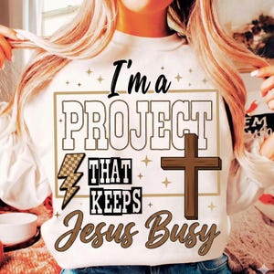 Könnte beinhalten: Weißes Sweatshirt mit dem Text "I'm a PROJECT THAT KEEPS Jesus Busy" in Schwarz und Braun. Das Design beinhaltet ein Kreuz, einen Blitz und Sterne. Ein lässiges Kleidungsstück mit religiösem Thema.