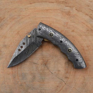 May include: A Damascus steel folding knife with a patterned blade and handle. The handle features brass accents and black rivets. The blade has three circular cutouts. The knife is open, displaying the full blade length.