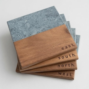 May include: A stack of four square coasters, each featuring a split design of grey stone and brown wood. The wooden portion of each coaster is engraved with one of the cardinal directions: east, south, north, or west, in a simple black font.