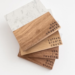 May include: A set of four square coasters featuring a split design of white marble and light brown wood. Each wooden section is engraved with black text: one more chapter, coffee kindle couch, shh. i'm reading, and plot over plans.